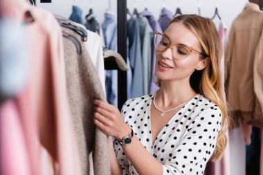 smiling, stylish businesswoman near clothes on hangers in showroom, blurred foreground clipart