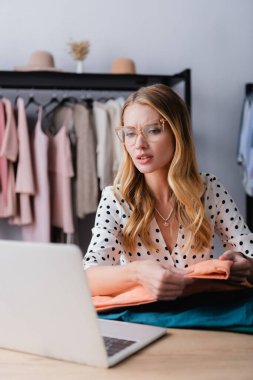 Düşünceli bir galeri sahibi elinde tekstil örnekleri tutarken dizüstü bilgisayara bakıyor, ön plan bulanık