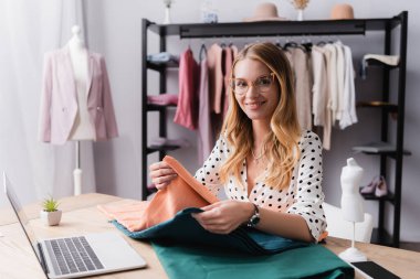 Gülümseyen galeri sahibi dizüstü bilgisayarın yanındaki kumaş örneklerini karşılaştırırken kameraya bakıyor.