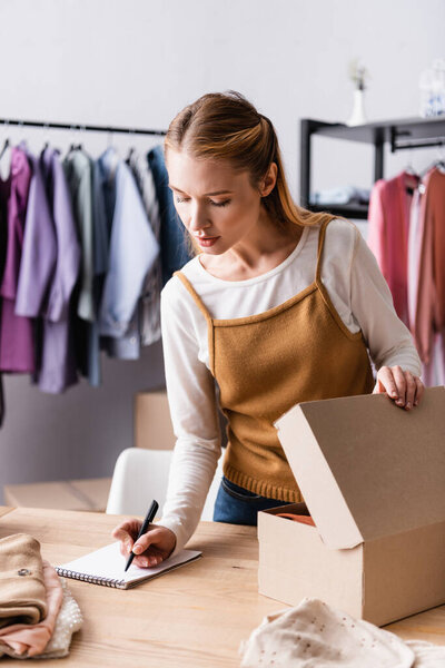 young seller writing order in notebook near carton package in clothes showroom