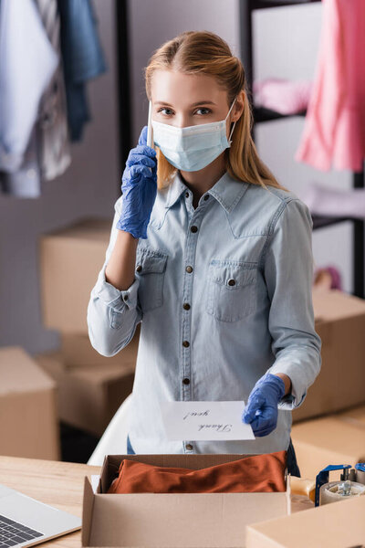 seller in medical mask, talking on smartphone while holding card with thank you lettering in showroom, blurred foreground