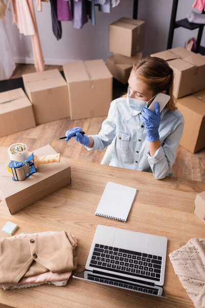 high angle view of showroom owner in medical mask, talking on smartphone near laptop