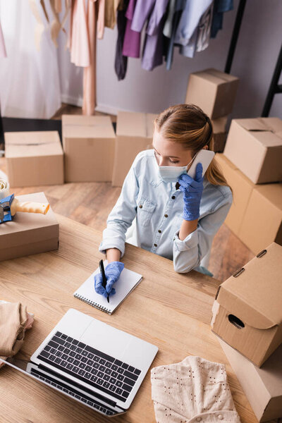high angle view of showroom owner in medical mask, writing in notebook while confirming order on mobile phone