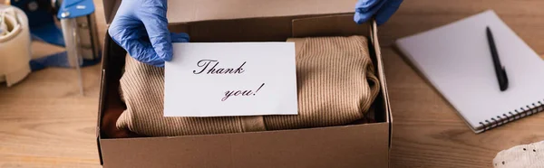 cropped view of seller in latex gloves holding card with thank you lettering near box with clothes, banner