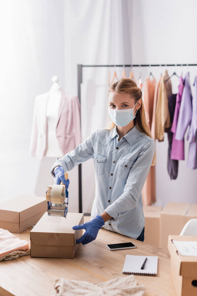 showroom owner in medical mask, looking at camera while sealing box with adhesive tape