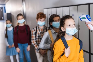 Tıbbi maskeli öğrencilere kızılötesi termometre kullanan öğretmen, bulanık arka planda çocukların yanında. 