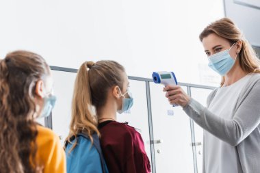 Teacher in protective mask using infrared thermometer on schoolgirls during quarantine in school  clipart