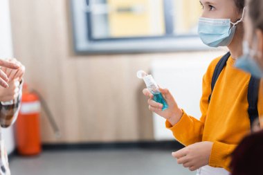 Tıp maskeli kız, koridordaki bulanık ön planda arkadaşlarının yanında el dezenfektanı tutuyor. 