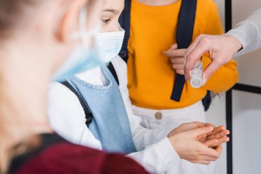 Öğretmen, çocukların yanında sağlık maskesi takan öğrencinin ellerine dezenfektan döküyor. 