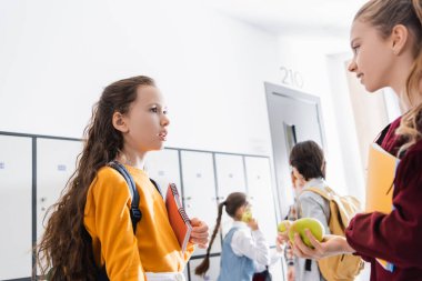 Okul koridorunda konuşan elmalı ve defterli kızlar. 