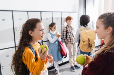 Elma ve defterle gülümseyen liseli kız bulanık arka planda sınıf arkadaşlarıyla konuşuyor. 