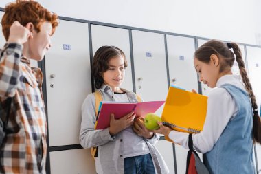 Sınıf arkadaşlarının yanında elmayı tutarken kitapçığa bakan kız 