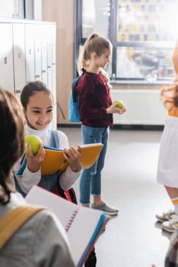 Elmalı ve defterli neşeli okul çocuğu koridordaki bulanık ön planda sınıf arkadaşlarına bakıyor. 