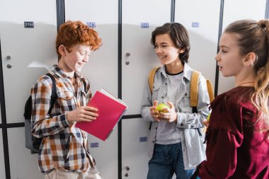 Okulda dolapların yanında duran defter, elma ve sırt çantalı neşeli çocuklar. 