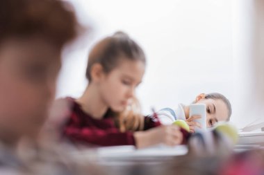 Sınıf arkadaşlarının yanında akıllı telefon kullanan okul çocuğu sınıfta bulanık ön planda 