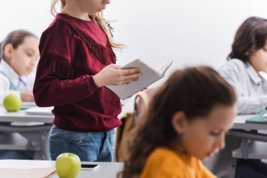 Akıllı telefon, elma ve sınıf arkadaşlarının yanında ders sırasında bulanık arkaplan üzerine kitap tutan okul çocuğu 