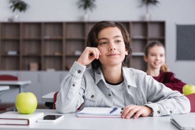 Gülümseyen öğrenci masasında elma ve defter olan cihazların yanından uzağa bakıyor. 