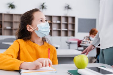 Tıp maskeli kız sınıfta defter ve elmanın yanındaki öğretmene bakıyor.