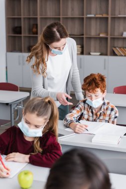 Tıp maskeli öğretmen ders sırasında not defterine parmağıyla işaret ediyor.