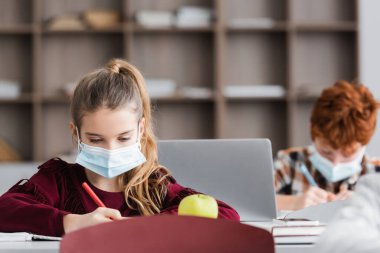 Koruyucu maskeli kız, bulanık ön planda elmanın yanında yazıyor.