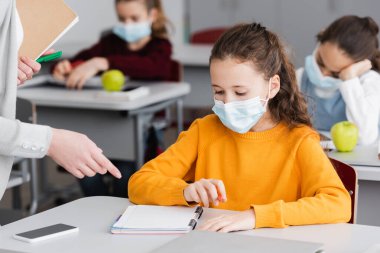 Öğretmen, tıp maskeli okul kızının yanındaki defteri işaret ediyor.