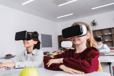 smiling classmates in vr headsets near pupils in classroom on blurred background clipart