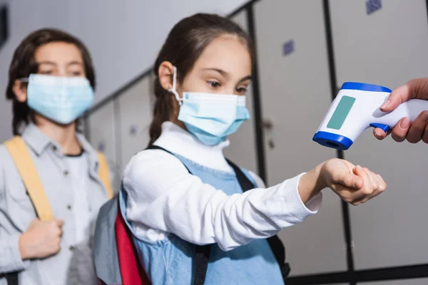 Teacher with non contact thermometer measuring temperature of schoolgirl in medical mask