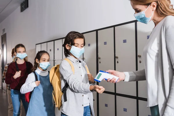 Schoolboy in medical mask standing near teacher with non contact thermometer in school corridor