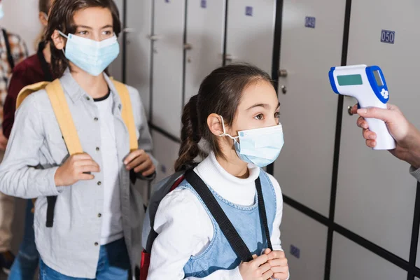Schoolkid in medical mask standing near teacher measuring temperature in school corridor