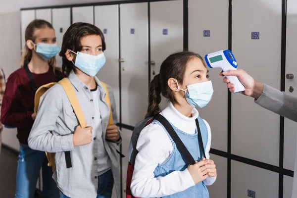 Teacher with non contact thermometer measuring temperature of schoolkid in medical mask near pupils