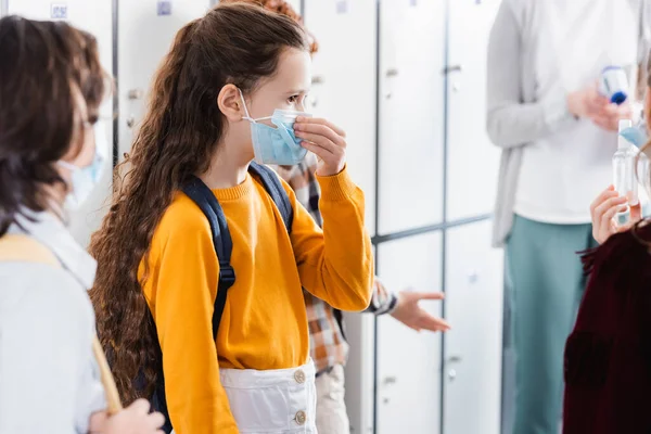 Schoolgirl wearing medical mask near friends with hand sanitizer on blurred foreground
