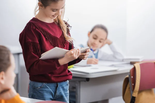 Uśmiechnięty Dzieciak Trzymający Książkę Pobliżu Uczniów Niewyraźnym Tle Klasie — Zdjęcie stockowe