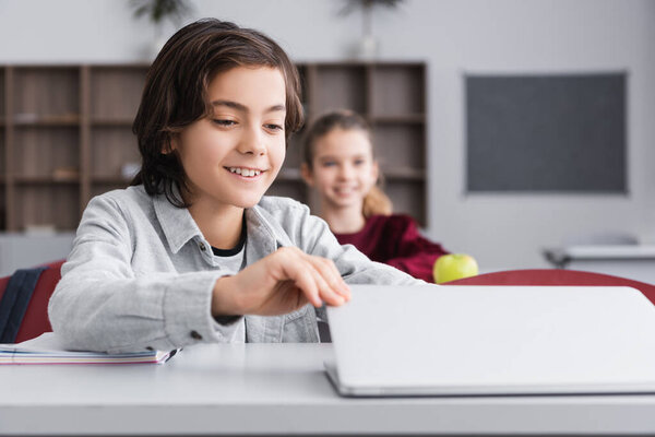 Cheerful schoolkid opening laptop near notebook on desk 