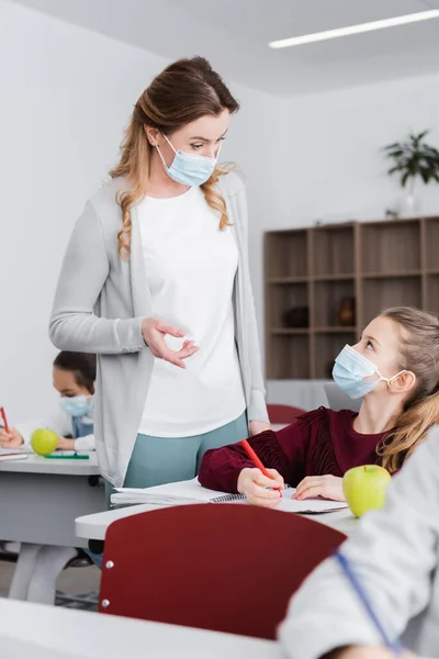 teacher in medical mask pointing with finger near schoolgirl in classroom