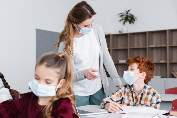 учитель в медицинской маске указывает пальцем на школьника в классе