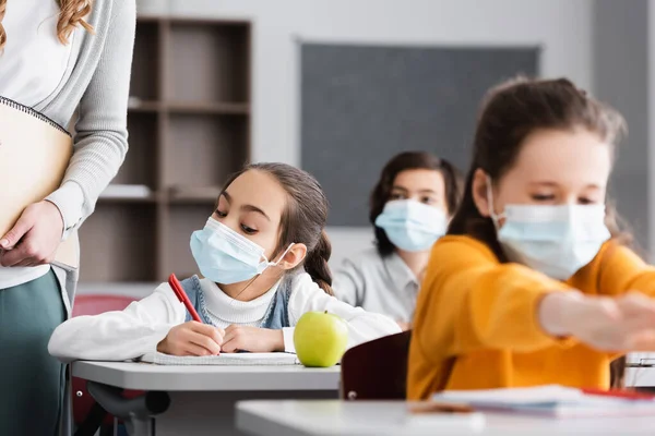 schoolgirl in medical mask near teacher, apple and classmates on blurred foreground