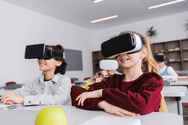 happy schoolchildren gaming in vr headsets during lesson, blurred background