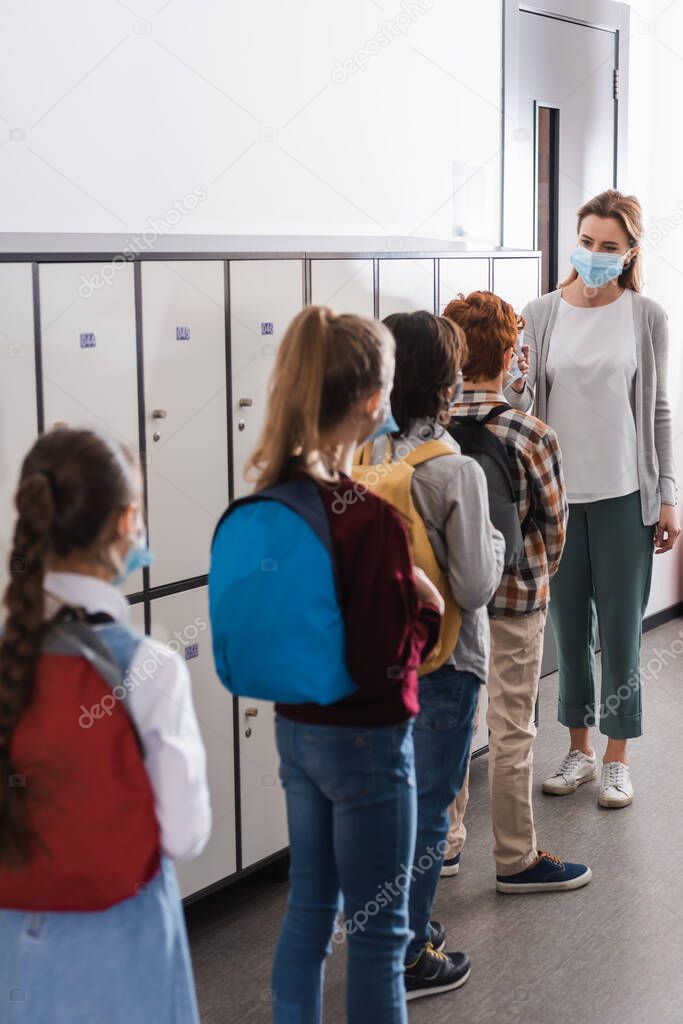 Teacher holding non contact thermometer near pupils in medical masks on blurred foreground in corridor 