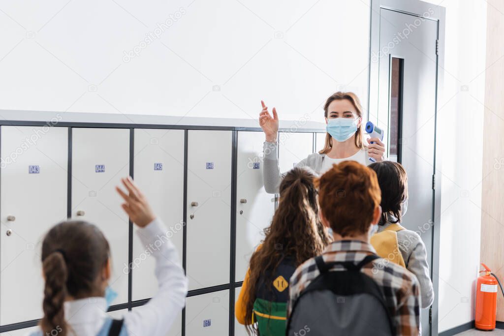 Teacher in medical mask holding non contact thermometer near schoolchildren on blurred foreground in corridor 