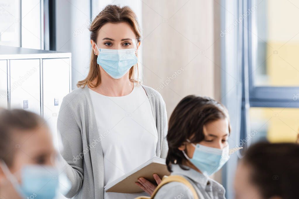 Teacher in protective mask holding notebook near pupils on blurred foreground 