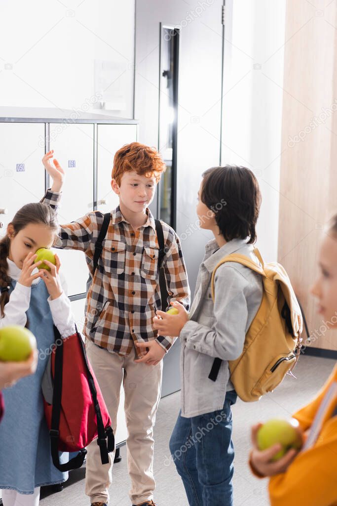 Schoolboy talking to friend with apple near friends on blurred foreground in school corridor 