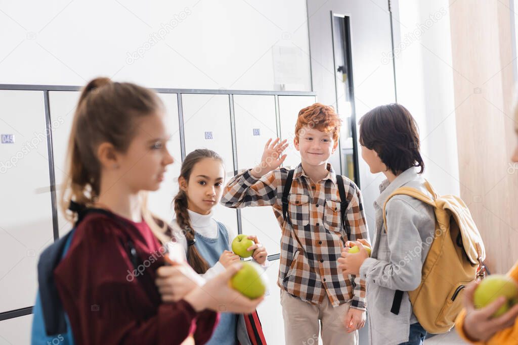 Smiling schoolboy waving hand near friends with apples on blurred foreground in school hall 