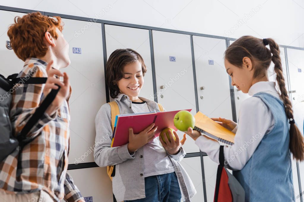 Smiling schoolboy looking at notebook near friends on blurred foreground and lockers in hall 