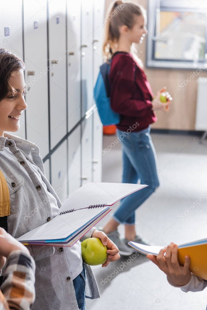 Smiling schoolkid with apple and notebook standing near lockers and classmates on blurred foreground 
