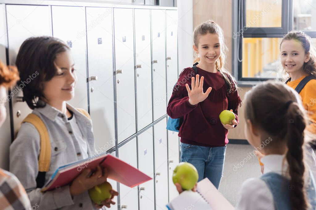 Smiling schoolgirl with apple waving hand to classmates in school corridor 
