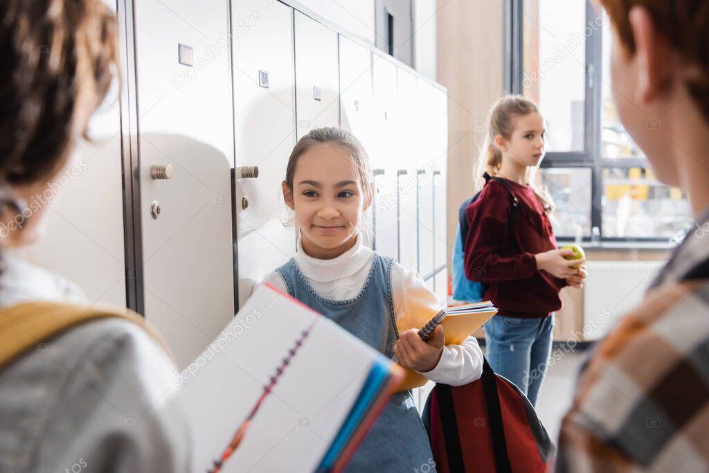 Schoolgirl with notebook smiling at friends on blurred foreground in school hall 