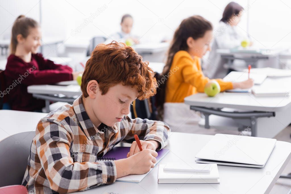 Schoolboy writing on notebook near laptop and smartphone on desk in classroom 