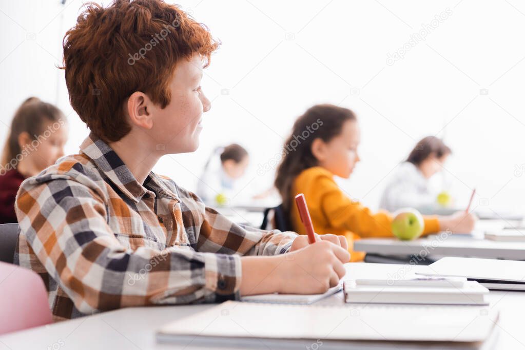 Schoolboy writing on notebook near laptop on blurred foreground in classroom 