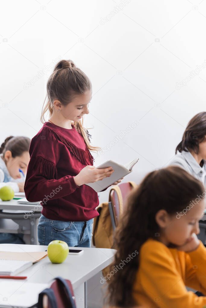 Cheerful schoolgirl reading book near classmates in classroom 