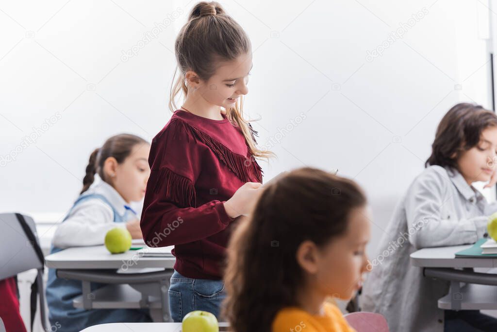 Smiling child standing near apple on desk and classmates on blurred background in school 